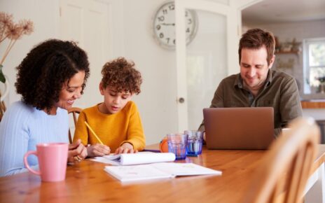 Father Works On Laptop As Mother Helps Son With Homework On Kitchen Table