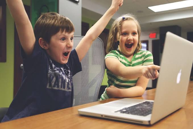 Two excited children celebrating while using a laptop together at a table.